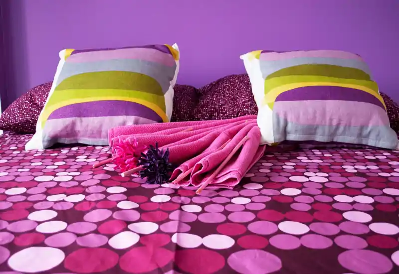 Imagen de dormitorio brillante y colorido centrada en una cama bien hecha con ropa de cama estampada, dos cojines decorativos y toallas dobladas — un espacio acogedor.