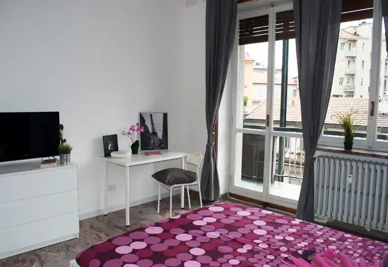 Dormitorio luminoso y bien decorado con colcha morada estampada, pequeño escritorio y silla, cómoda con TV y gran ventana/puerta al balcón que aporta mucha luz natural.
