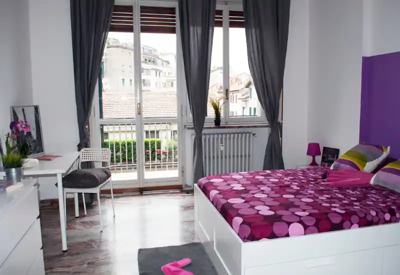 Dormitorio moderno y luminoso con estructura de cama blanca y ropa de cama morado/rosa vibrante, gran ventana/puerta a un pequeño balcón que aporta mucha luz natural. Escritorio compacto, plantas y decoración coordinada crean un ambiente acogedor y bien cuidado, ideal para una persona o pareja.