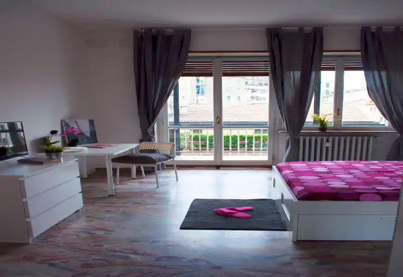 Dormitorio moderno y luminoso con una cama con estampado junto a una gran ventana y balcón. La habitación dispone de un escritorio pequeño, cómoda y textiles acogedores, ideal para una estancia cómoda.