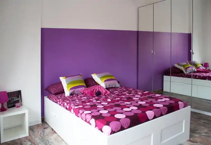 Dormitorio moderno y luminoso con cama doble estampada, cojines decorativos y armario con espejo que añade profundidad. La pared morada y los textiles coordinados crean un espacio acogedor y con estilo.