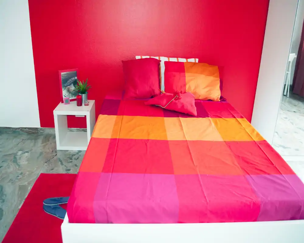 Bright, colorful bedroom with a large bed dressed in vibrant red and orange linens, a small bedside table with decor and a minimalist floor. The strong color palette and natural light create an inviting, modern sleeping space.