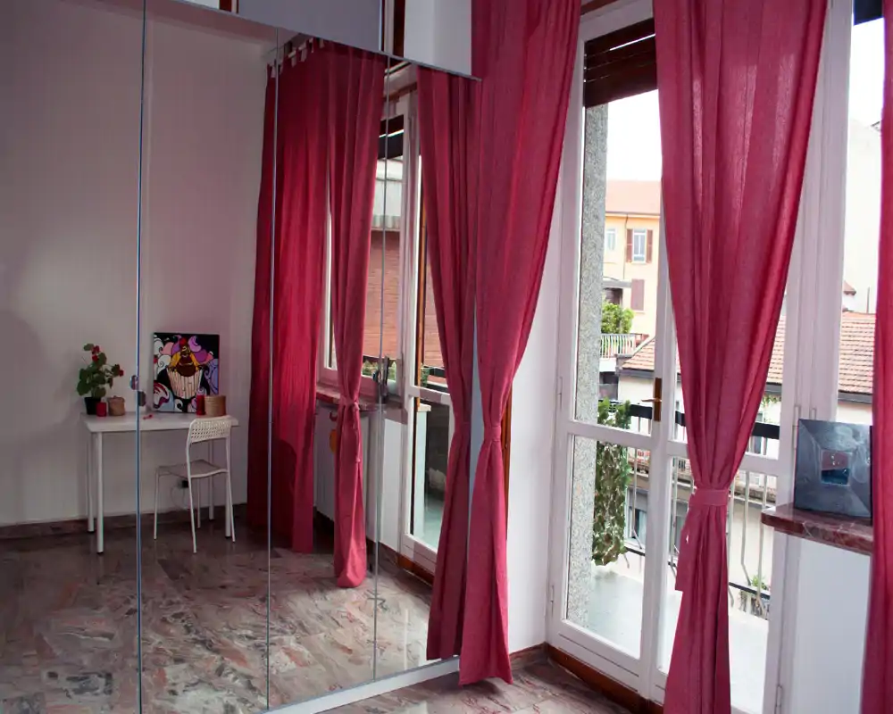 Empty bedroom-like space with large mirrored wardrobe and deep red curtains; bright natural light from floor-to-ceiling windows and a small desk by the wall.