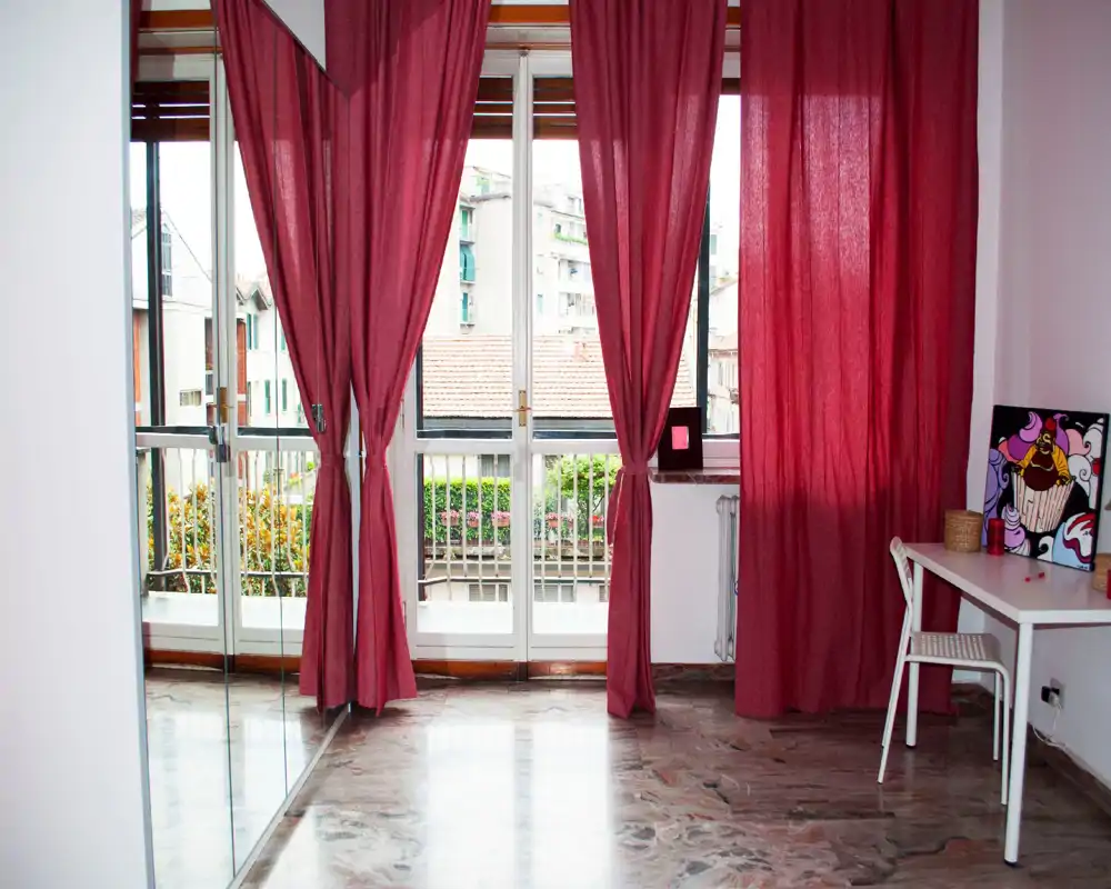 Bright living area with large floor-to-ceiling windows and burgundy curtains, a small desk and decorative artwork create a cosy corner with polished marble flooring and a balcony view.