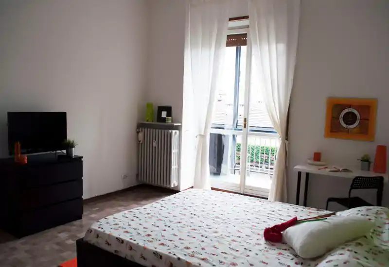 Chambre lumineuse et aérée avec un grand lit à motifs face à une porte-fenêtre vers le balcon. Mobilier moderne et minimaliste, petit bureau, commode TV et lumière naturelle douce à travers les voilages.