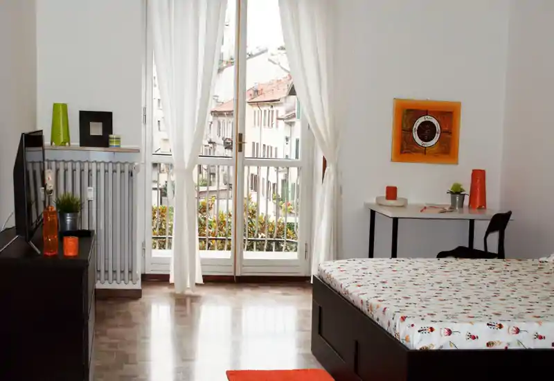 Camera da letto luminosa e ben arredata con letto matrimoniale, scrivania e ampie porte sul balcone che illuminano l'ambiente — dettagli colorati e pavimento in parquet valorizzano l'annuncio.