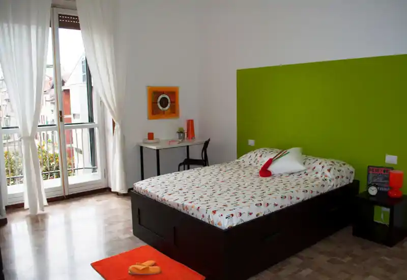 Chambre moderne et lumineuse avec lit double, tête de lit verte et grande fenêtre laissant entrer la lumière naturelle ; bureau et table de chevet simples.