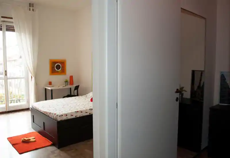 Chambre lumineuse et sobrement meublée avec un lit double, un tapis près du lit et un petit bureau près du balcon. La pièce est inondée de lumière naturelle par la porte-fenêtre et les voilages.