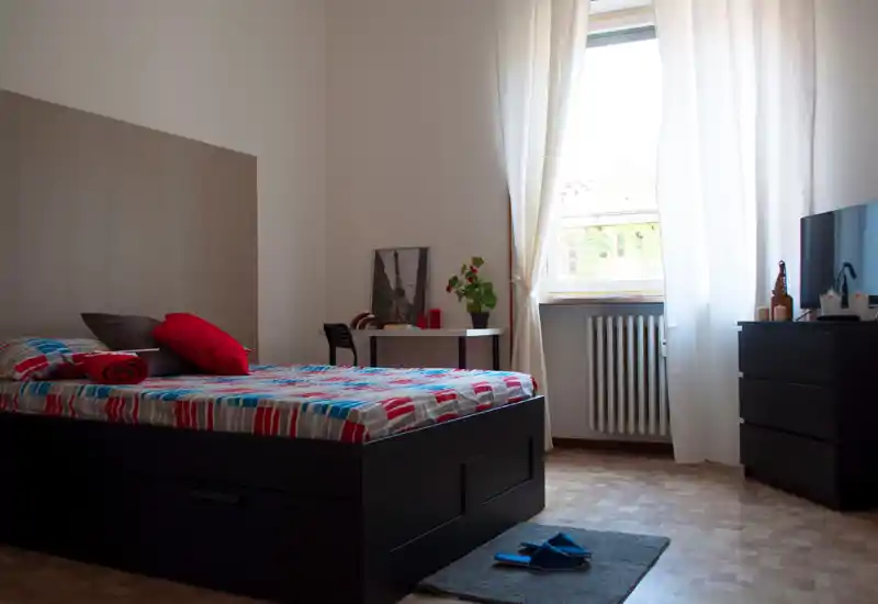 Chambre lumineuse et bien rangée avec un grand lit, une literie colorée, une table d'appoint et une lumière naturelle provenant de la fenêtre. Mobilier minimaliste et touches chaleureuses pour une ambiance accueillante.