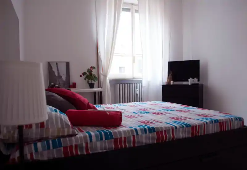 Chambre lumineuse et soignée avec un grand lit recouvert d’une couette rayée colorée, lumière naturelle douce venant de la fenêtre et quelques éléments décoratifs simples (coussins, table de chevet).