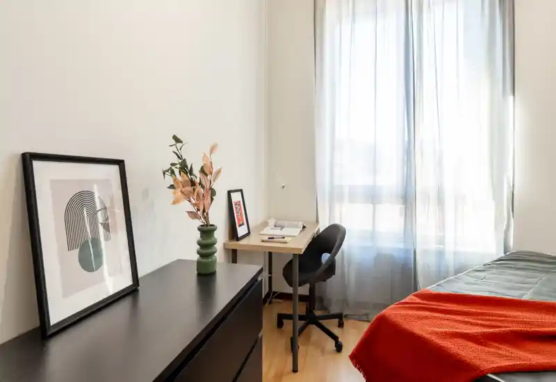 Bright, modern bedroom with a visible bed corner, a compact desk by the window and tasteful decorative touches including a framed print and a cozy red throw.