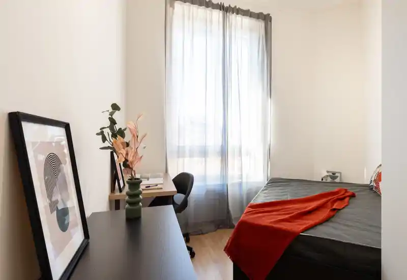 Bright, well-styled bedroom with a neatly made bed, soft natural light from a large window, and a small desk area — ideal for restful stays or a work-and-sleep setup.