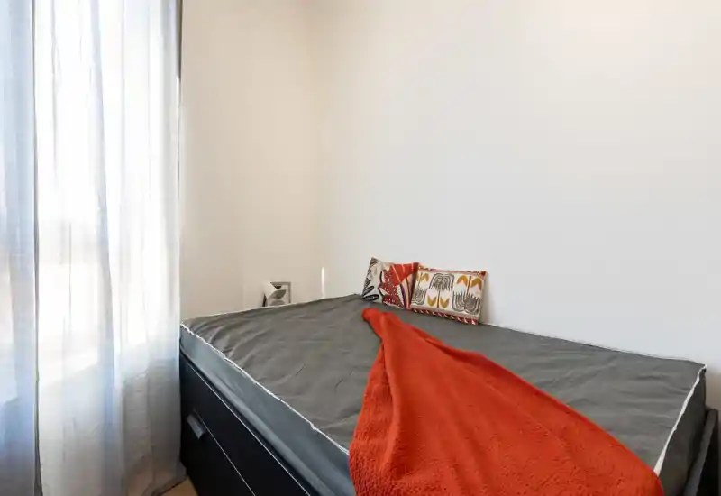 Bright, minimal bedroom with a neatly made single bed, decorative pillows and a warm orange throw beside a sheer-window letting in soft daylight.