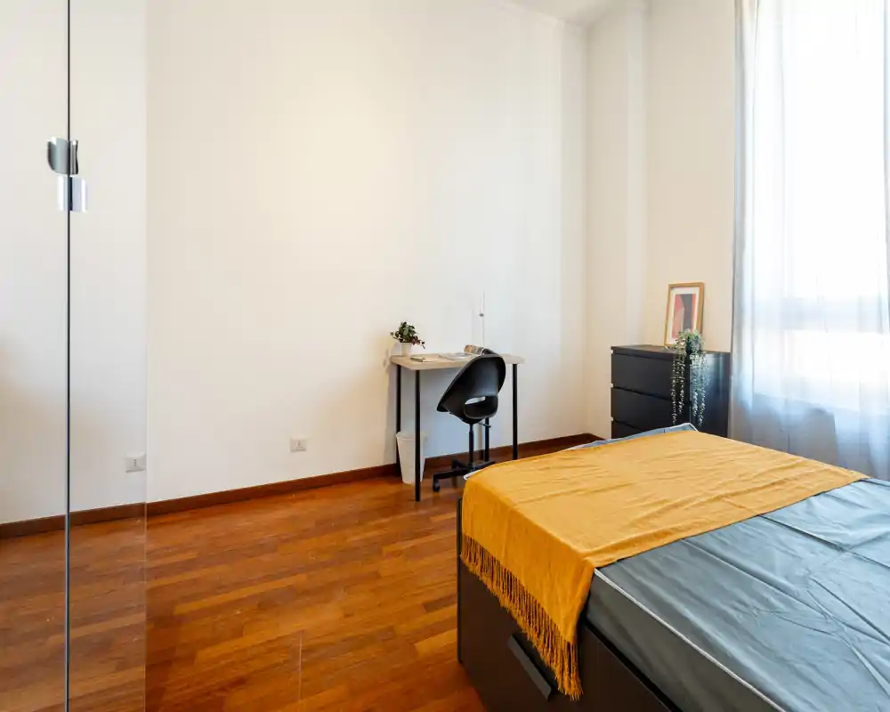 Dormitorio amplio y luminoso con cama bien arreglada y manta mostaza, un pequeño espacio de trabajo y luz natural de una ventana grande.