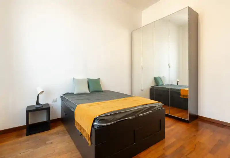 Dormitorio individual moderno y luminoso con cama-baúl negra elevada, armario con puertas espejo y suelo de madera cálido. Estilo minimalista limpio con cojines y una manta amarilla que aportan confort.