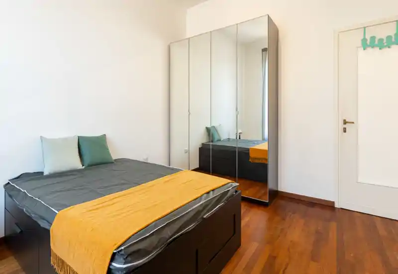 Dormitorio luminoso y minimalista con cama doble bien hecha, cojines y una manta amarilla; armario con espejos que aporta profundidad.