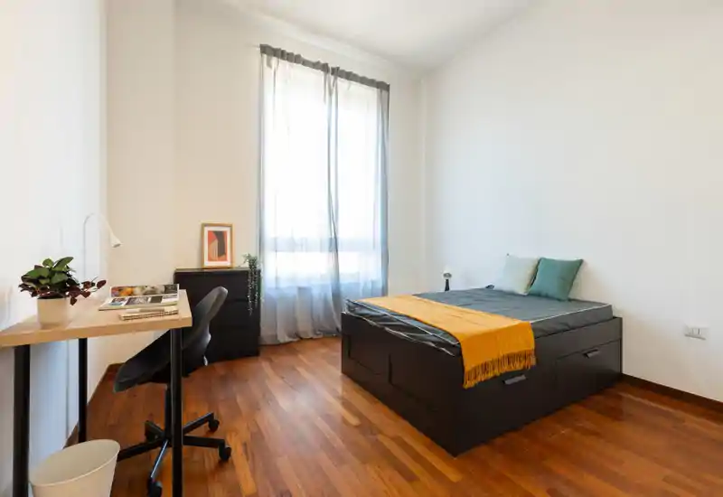 Dormitorio luminoso y minimalista con cama doble con cajones, cojines y una manta cálida. Escritorio junto a la ventana y cómoda pequeña generan un espacio funcional y aireado con suelo de madera.