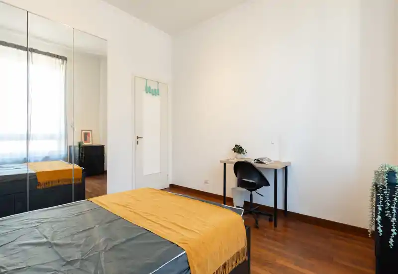 Dormitorio moderno y luminoso con cama doble bien hecha, una manta amarilla cálida y un pequeño escritorio junto a la pared. Armario con espejos y suelo de madera que amplían el espacio.