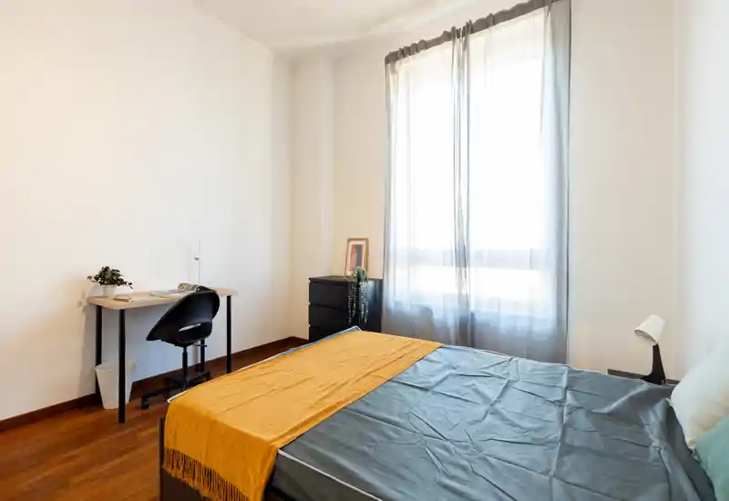 Dormitorio luminoso y minimalista con cama doble de ropa oscura y una manta mostaza, un pequeño escritorio y una gran ventana que aporta luz natural; perfecto para estancias cómodas y teletrabajo.