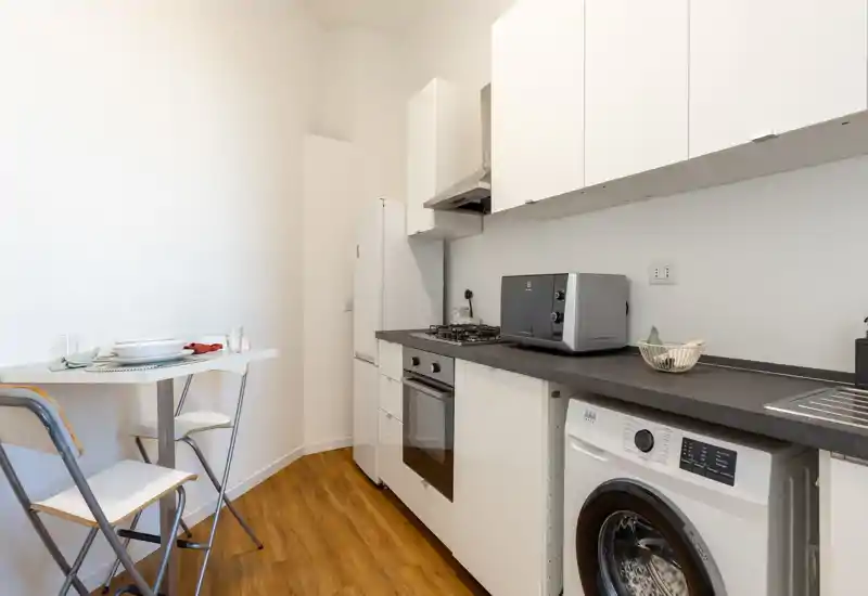 Compact modern kitchenette with white cabinets, built-in oven, washing machine and a small dining nook — bright, clean and functional for city stays.