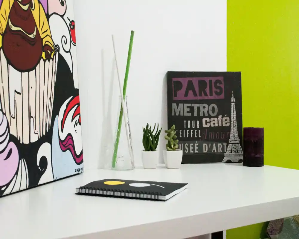 Rincón decorativo moderno y luminoso con un cuadro de París, pequeñas suculentas en maceta y un cuaderno sobre una mesa blanca — una esquina con estilo para salón o espacio de trabajo.