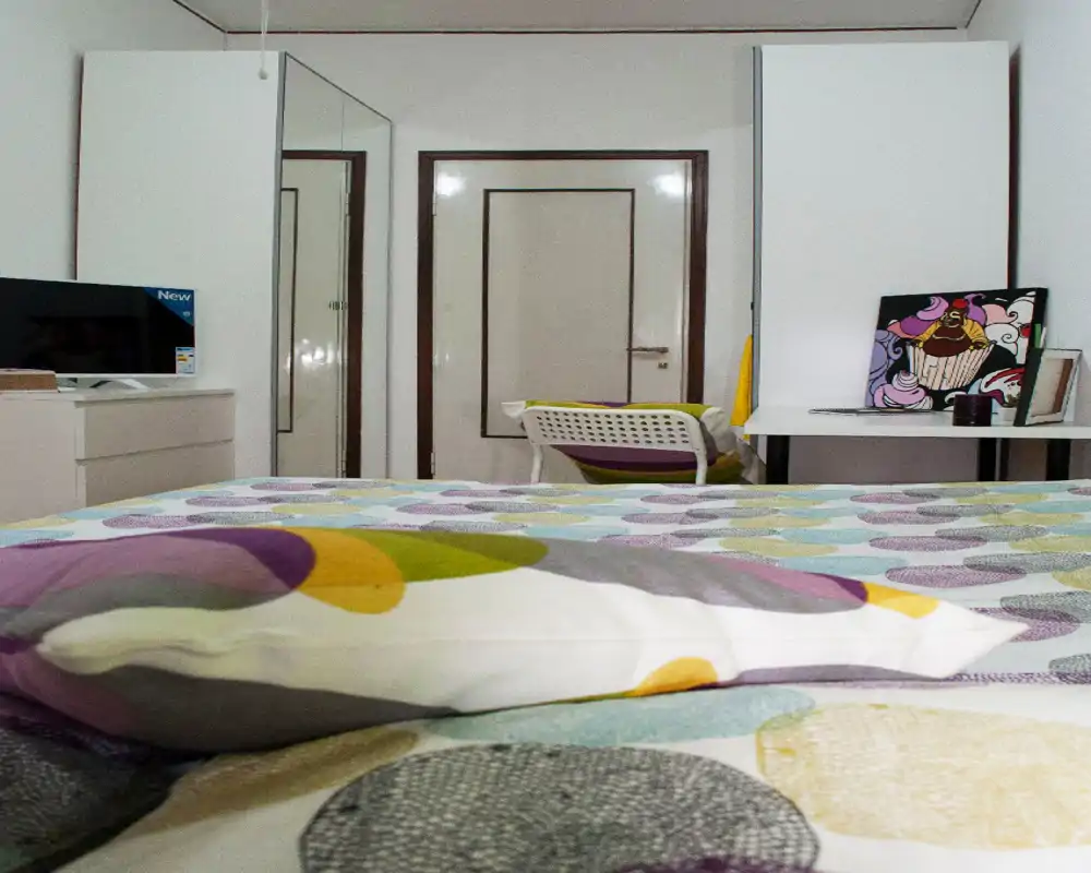 Dormitorio ordenado y luminoso centrado en una colcha con estampado, con escritorio y armario al fondo; decoración colorida adecuada para un anuncio.