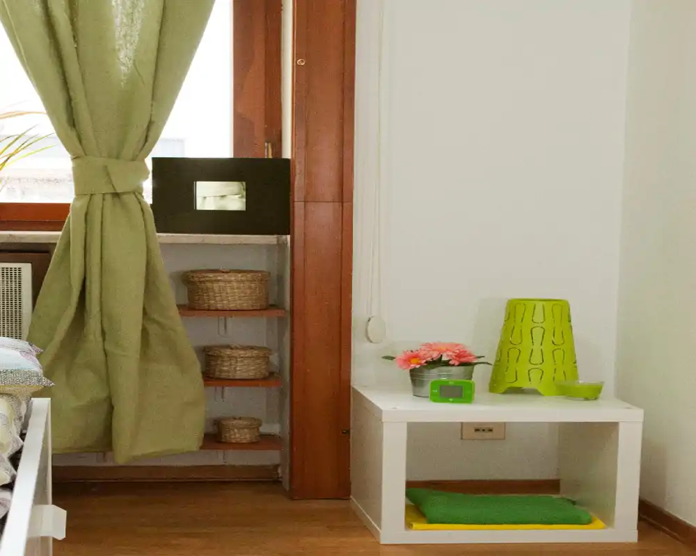 Rincón de dormitorio pequeño y ordenado con cortina verde, mesita y estantería con cestas; la luz natural ilumina el espacio.