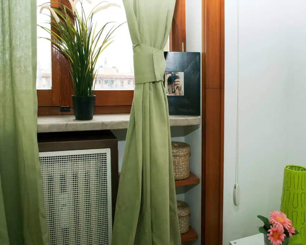 Rincón de ventana luminoso con cortinas verdes, planta en el alféizar, cestas de mimbre en pequeños estantes y cubierta del radiador debajo; ideal para destacar la luz natural y los detalles decorativos.