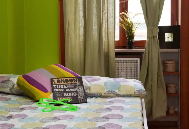 Dormitorio luminoso y colorido con ropa de cama estampada, cojines decorativos y luz natural de la ventana; ordenado y acogedor.