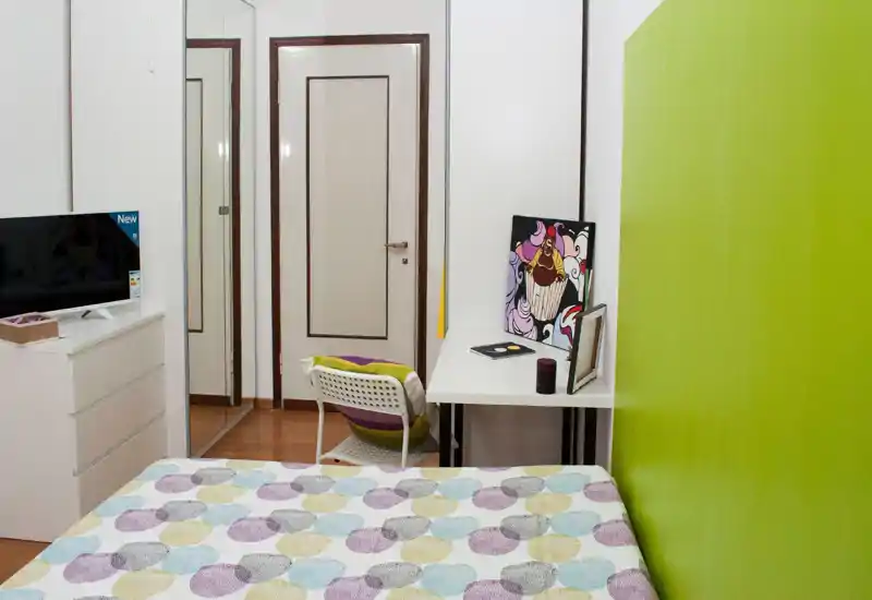 Dormitorio compacto y luminoso con colcha de diseño, escritorio y silla junto a una pared verde intensa, y un armario con espejo que aporta almacenamiento y sensación de amplitud.