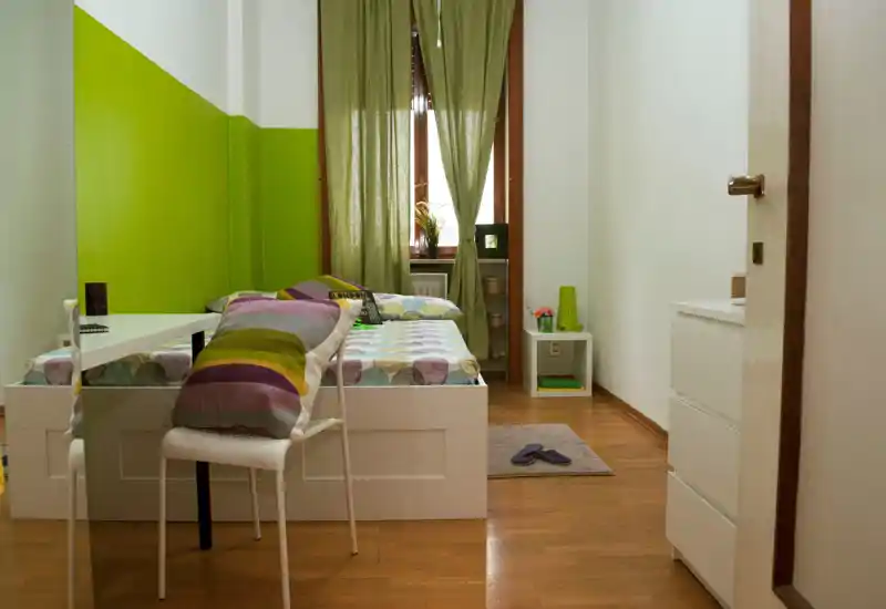 Dormitorio individual ordenado con cama blanca, ropa de cama estampada y detalles verdes; una ventana con cortinas verde claro aporta luz natural y una mesita de noche pequeña.