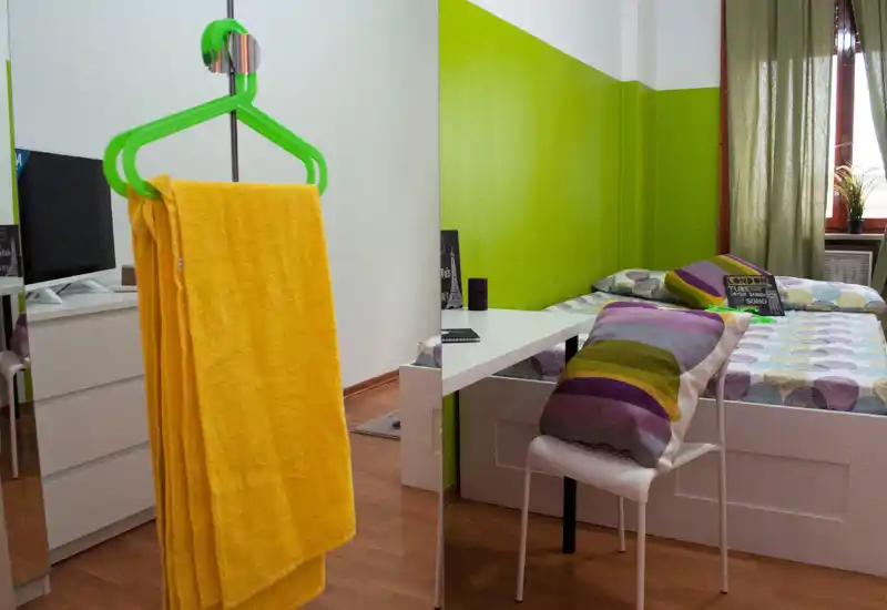 Dormitorio luminoso y colorido con cama doble, ropa de cama estampada y cojines decorativos; un pequeño escritorio junto a la cama y una ventana que aporta luz natural.