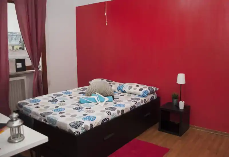 Dormitorio luminoso y bien decorado con cama doble, ropa de cama estampada, mesita y lámpara. La pared roja y el suelo de madera crean un ambiente cálido y moderno.
