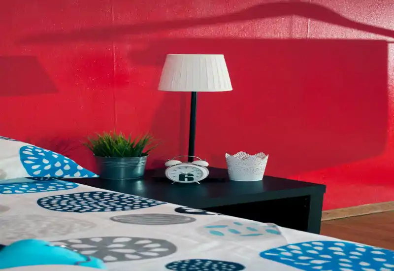 Rincón de dormitorio luminoso y estilizado con ropa de cama estampada, una mesilla con lámpara, despertador y planta frente a una pared roja intensa — perfecto para destacar un cabecero acogedor.