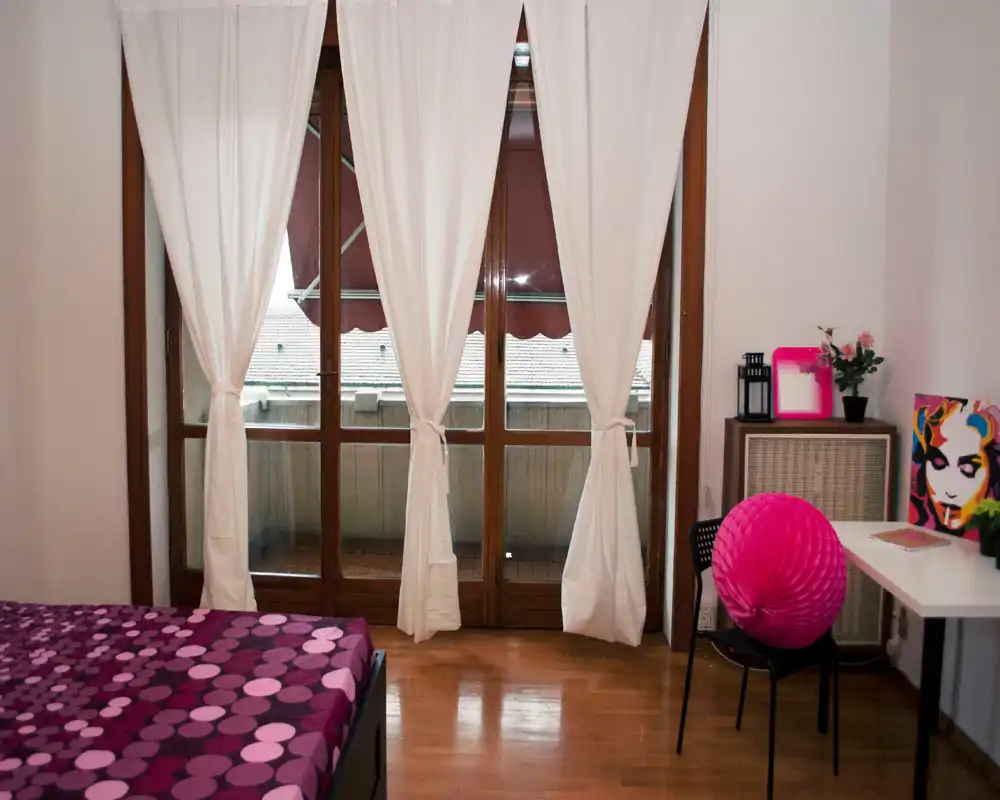 Dormitorio luminoso y con estilo: cama morada con estampado, grandes puertas al balcón con cortinas blancas y una pequeña zona de escritorio con decoración colorida — ideal para una estancia acogedora.