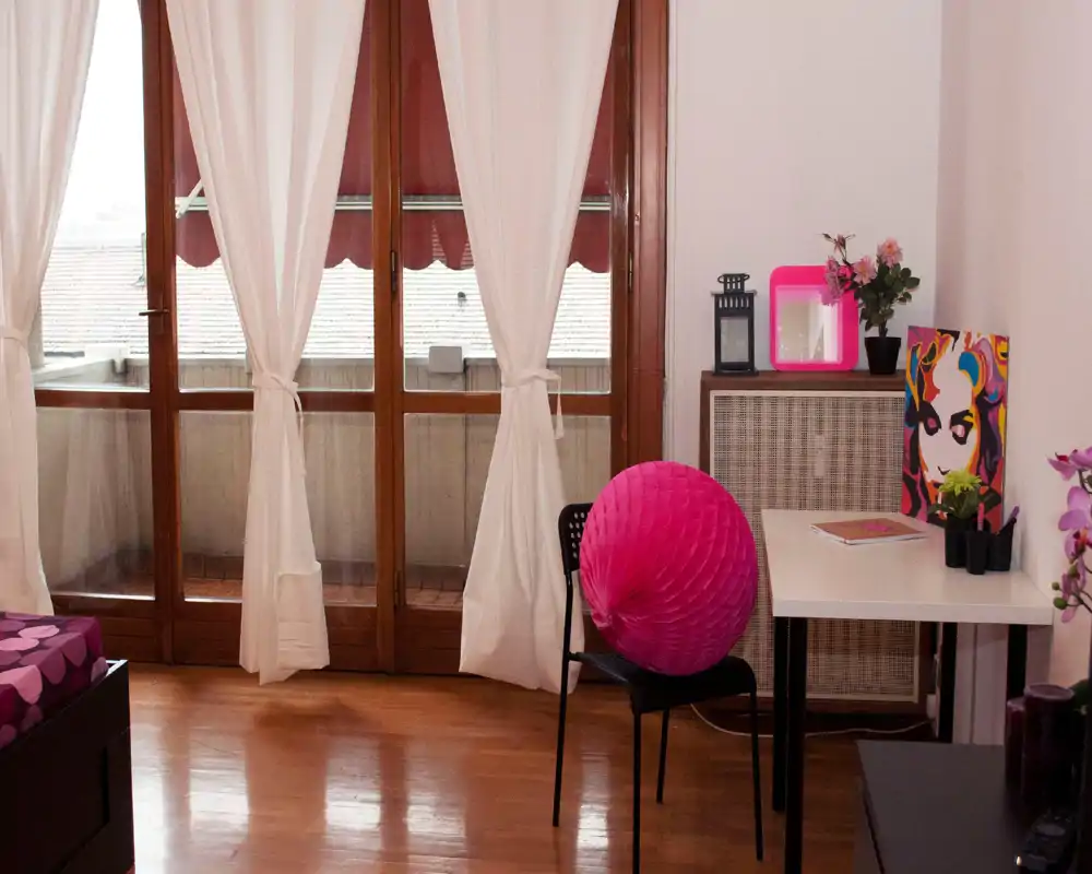 Rincón de dormitorio luminoso y cálido con suelo de madera, cama parcialmente visible a la izquierda, escritorio con elementos decorativos y puerta balcón con cortinas blancas que dejan pasar la luz natural.