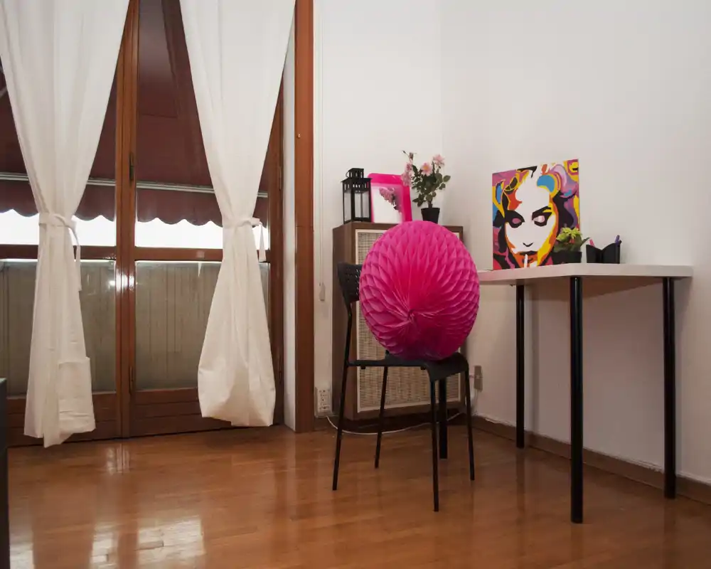 Pequeña zona de estar luminosa con suelo de madera, escritorio y silla, cuadros decorativos y una puerta balcón que aporta luz natural — espacio sencillo y ordenado ideal para un apartamento pequeño.