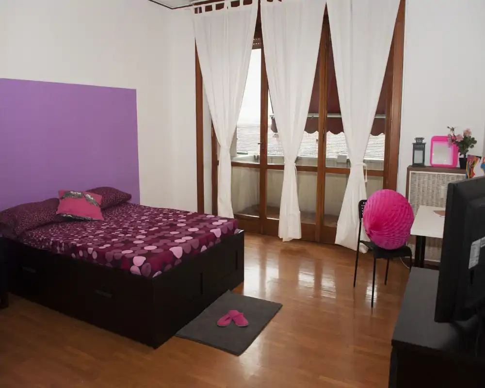 Dormitorio luminoso y bien decorado con cama doble con ropa de cama morada estampada, suelos de madera y grandes puertas al balcón que permiten la entrada de luz natural.