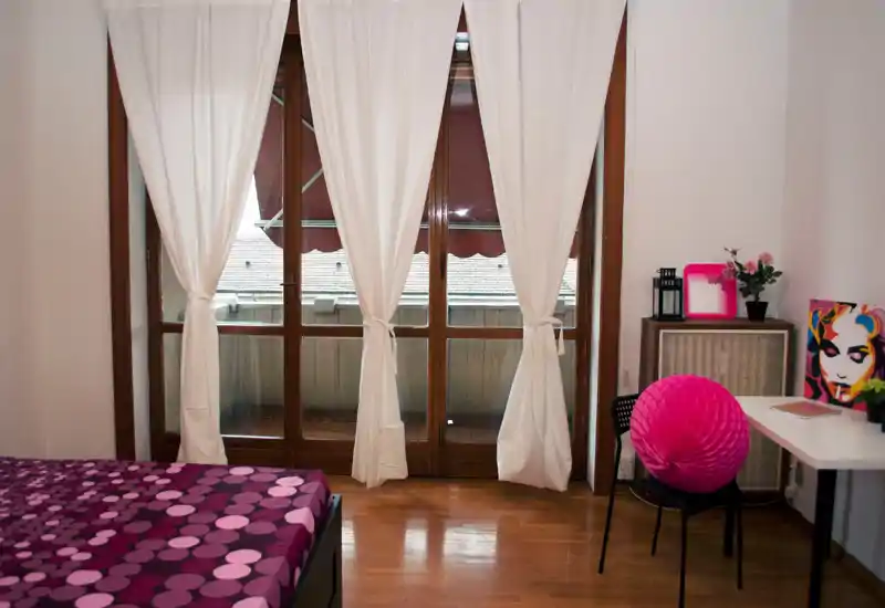 Dormitorio luminoso y con estilo: cama morada con estampado, grandes puertas al balcón con cortinas blancas y una pequeña zona de escritorio con decoración colorida — ideal para una estancia acogedora.