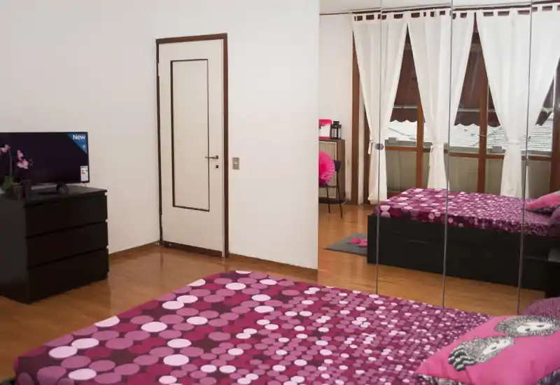 Dormitorio luminoso y acogedor con ropa de cama rosa estampada, suelo de madera y armario con espejo que refleja la luz natural.