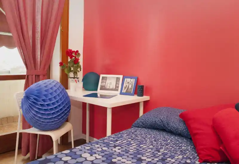 Rincón de dormitorio luminoso y colorido con cama vestida con ropa de cama azul estampada, cojines rojos y un pequeño escritorio blanco junto a la ventana con marcos y un jarrón.