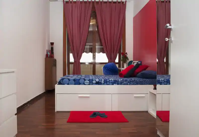 Dormitorio moderno y luminoso con cama plataforma, ropa de cama azul con estampado y detalles en rojo. La luz natural y la decoración coordinada crean un espacio acogedor y atractivo para anuncios.