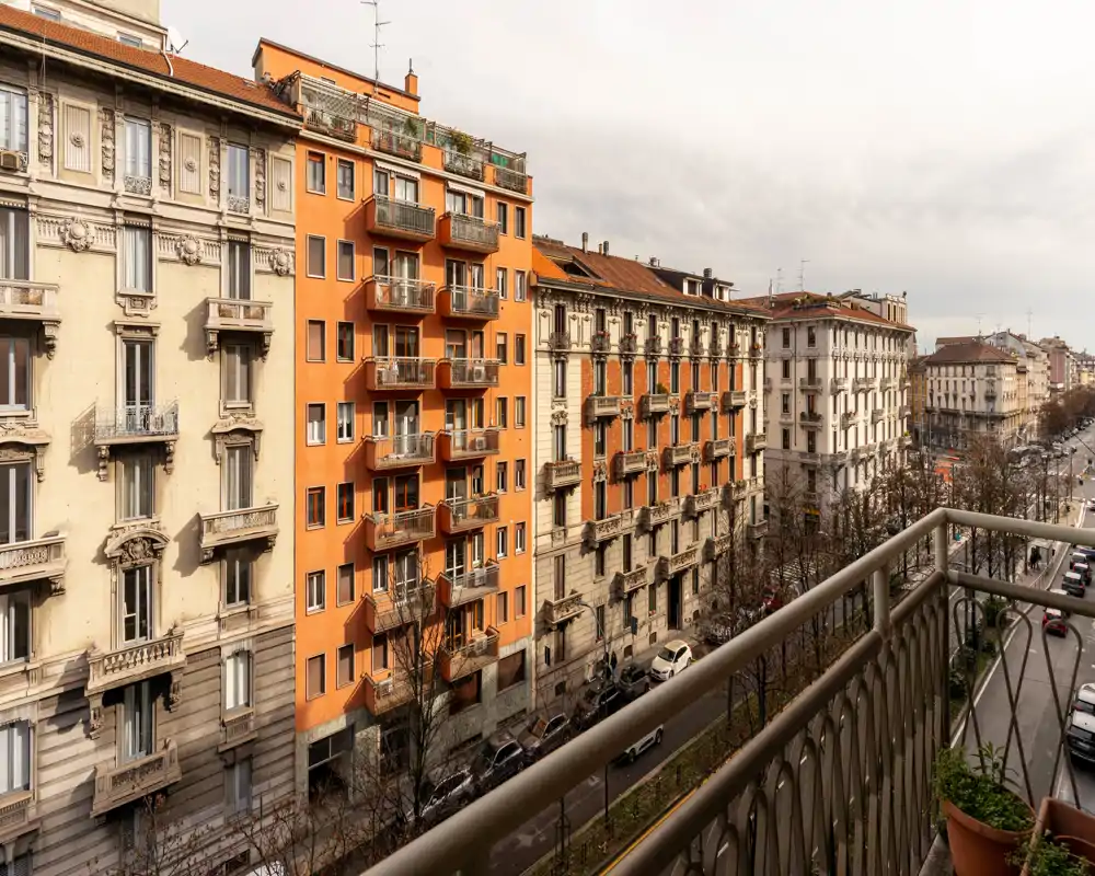 Balcón con vistas a una calle urbana y elegantes fachadas clásicas; la barandilla metálica y una maceta en primer plano enmarcan la escena.