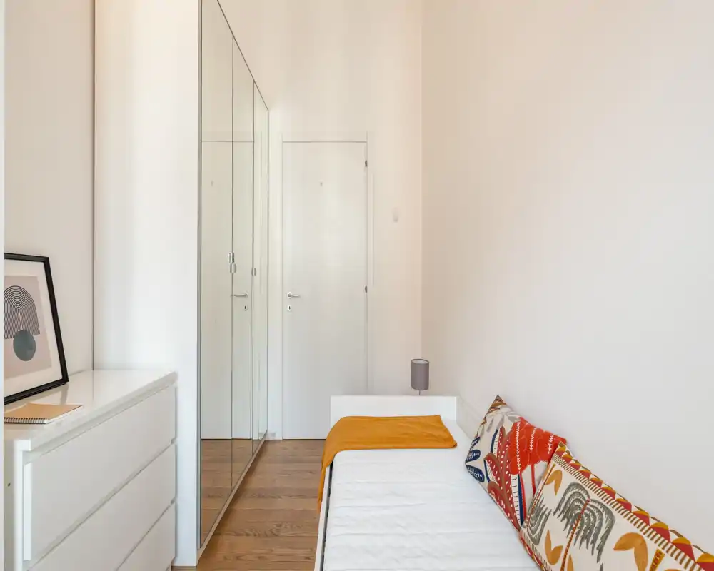Dormitorio individual luminoso con cama blanca compacta, cojines con estampado y suelo de madera cálida; armario con puertas espejo y cómoda sencilla que crean una sensación limpia y aireada.