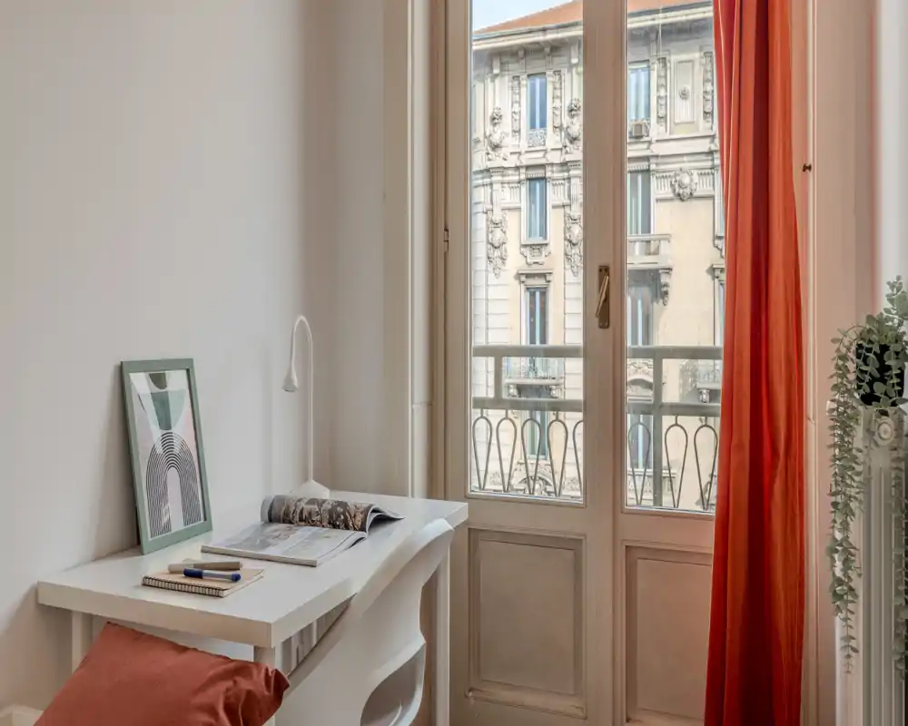 Rincón de dormitorio luminoso con escritorio blanco y silla junto a una ventana alta, cortina roja cálida y planta decorativa — ideal como espacio de trabajo tranquilo con vistas a la ciudad.