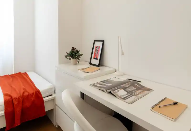 Angolo camera luminoso e minimal con una scrivania bianca e un letto con un caldo plaid rosso — ideale per un appartamento compatto e di design.