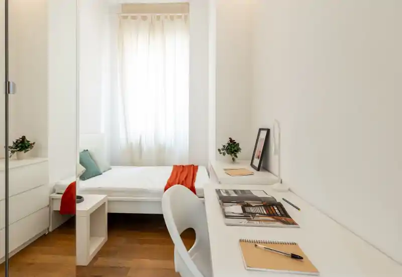 Camera da letto luminosa e minimal con letto singolo bianco, tessili accoglienti e area scrivania funzionale vicino alla finestra.