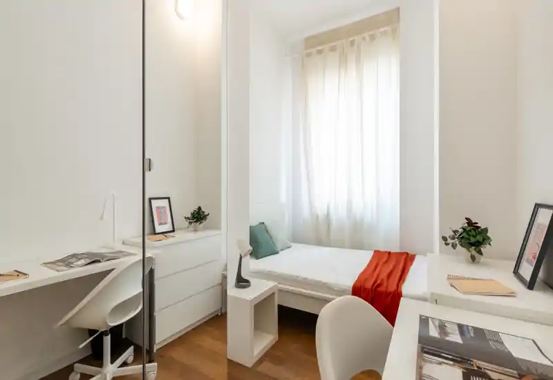 Camera da letto singola luminosa e minimal con letto bianco vicino alla finestra, un plaid rosso e una piccola scrivania. La luce naturale inonda l'ambiente, creando un'atmosfera moderna e rilassante.