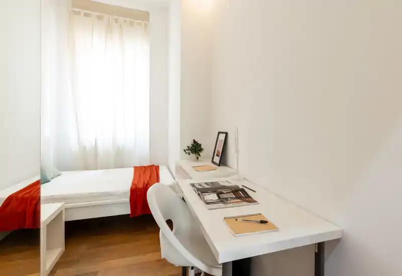 Dormitorio luminoso y minimalista con una cama individual junto a la ventana y un escritorio blanco perfecto para estudiar o teletrabajar. El suelo de madera y la manta roja añaden calidez.