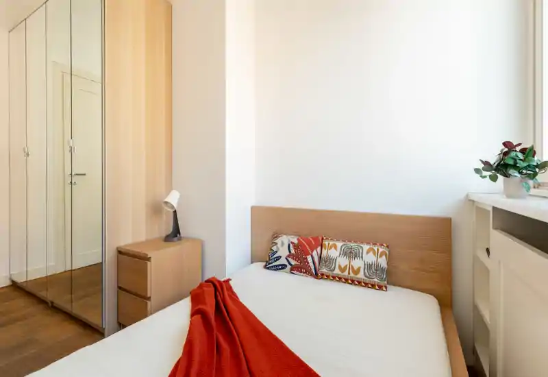 Camera da letto luminosa e minimale con letto in legno, cuscini colorati e plaid rosso, armadio con ante a specchio e comodino — uno spazio accogliente e ben illuminato.